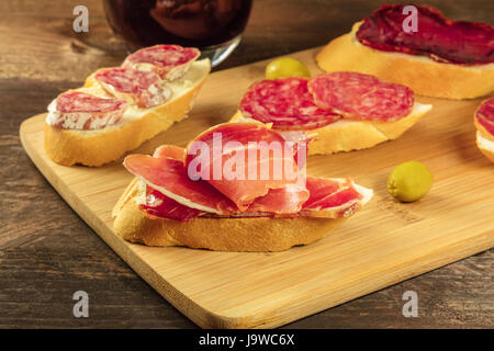 A photo of embutidos tapas, sandwiches with jamon, salchichon, lomo, and other Spanish sausages, with a glass of red wine and green olives, on a dark  Stock Photo