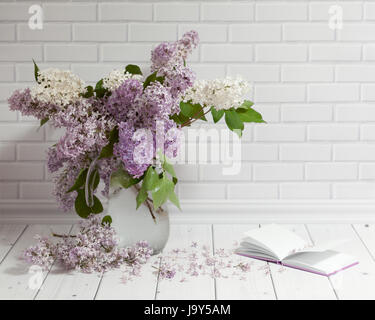 Beautiful bouquet of white and violet lilac flowers in glass vase with opened note-book on the white brick and wood background. Stock Photo