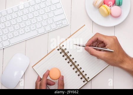 Man writing on open notebook on white desk with colorful macaroons, keyboard. Stock Photo