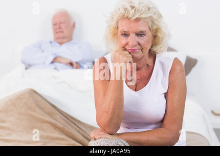 Discouraged old woman sitting on the bed with old man sleeping Stock Photo