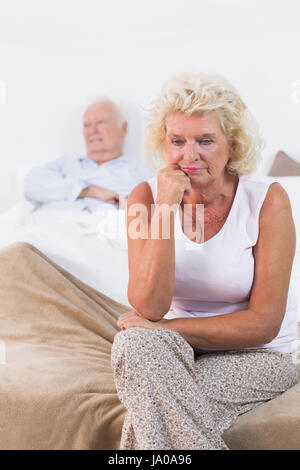 Discouraged old woman sitting on the bed with old man in background Stock Photo