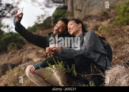 Young couple out on hike in mountains taking selfie with smart phone. Relaxed young man and woman sitting on country path and taking self portrait by Stock Photo