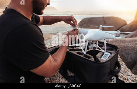 Young man sitting at rocky beach with a drone. Man setting up / dismantling a drone. Stock Photo