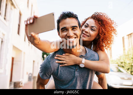 Smiling young woman riding piggy back on her partner while taking a selfie with her smartphone. Young man carrying his girlfriend on his back in a pla Stock Photo