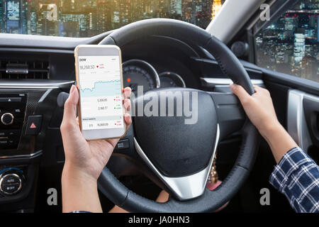 Business woman driver a touch screen of white smartphone check stock market or exchange and hand holding steering wheel in a car with night city backg Stock Photo