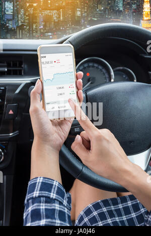 Business woman driver a touch screen of white smartphone check stock market or exchange and hand holding steering wheel in a car with night city backg Stock Photo