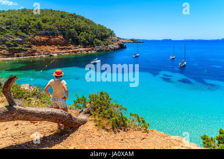 Young woman tourist standing on rock cliff edge and looking at beautiful Cala Salada bay famous for its azure crystal clear sea water, Ibiza island, S