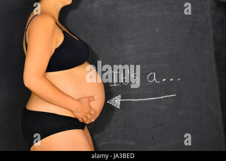 A pregnant woman is standing in front of a chalkboard with the words It's a... written on it and an arrow pointing to her tummy. Stock Photo