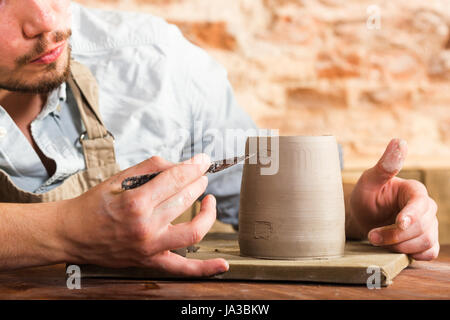pottery, stoneware, ceramics art concept - closeup on craftsman bent over a clay cup with tool, macro of male hands connecting pieces of fireclay, young male sits at a workshop behind the table. Stock Photo