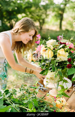 bouquet, people and floral arrangement concept - woman with beautiful bouquet of pink peonies, yellow and white roses, carnations and daisies, female florist at the table with flowers in garden. Stock Photo