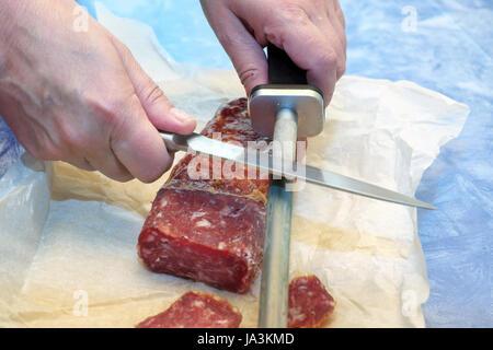 Female hands sharpening chef knife next to salami laid on a wrapping paper Stock Photo