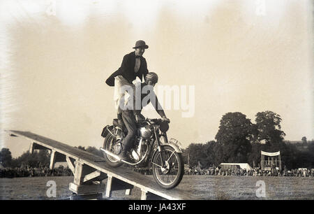 1950s, historical, clown stands on seat of a motocycle holding onto a the driver as it lands on a wooden ramp, England, UK. Stock Photo