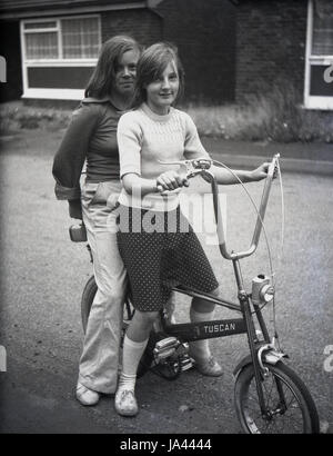 1970s, historical, two young girls stting outside on a Tuscan bike, a chopper type bicycle with high handlebars and different sized wheels, a popular and iconic bike of this era. Stock Photo