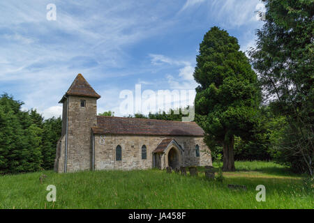 The Holy Trinity parish church in Godington in Oxfordshire. The church is surrounded by trees in a green pasture with a blue sky and wispy clouds. Stock Photo