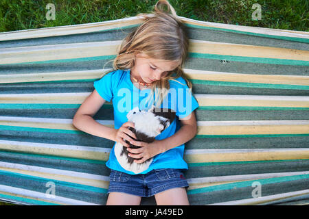 Child blond girl sitting or lying in a hammock and holding her guinea pig pet animal. Stock Photo