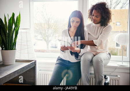 Two young businesswomen working on a tablet computer as they sit together on a bright windowsill in the office of their small business Stock Photo