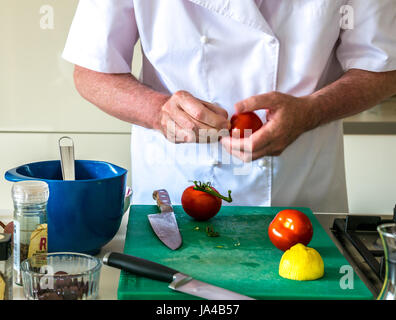 A man in chef whites preparing tomatoes on a green chopping board on a kitchen counter with other kitchen utensils and white cupboards in background Stock Photo