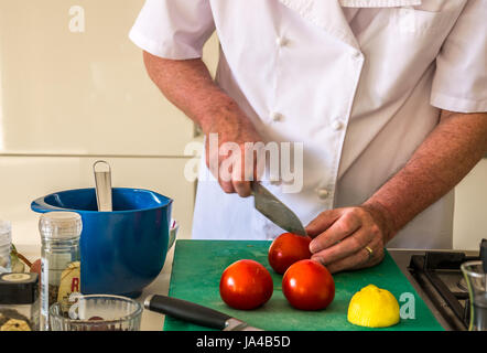 Older man in chef whites preparing tomatoes on chopping board on kitchen counter with kitchen utensils and white cupboards in the background Stock Photo