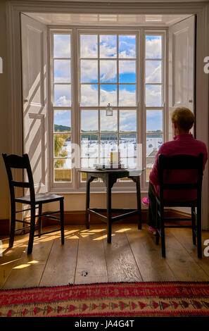 A woman sitting on a chair inside a cottage looking out through a large sunlit window to a view of boats moored in the harbour. Stock Photo
