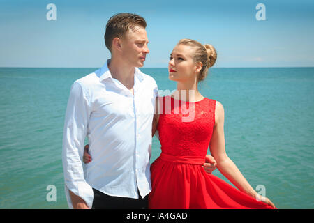 Man is hugging a woman delicatly posing with her by the sea. Portrait of a beautiful couple on vacation. Stock Photo