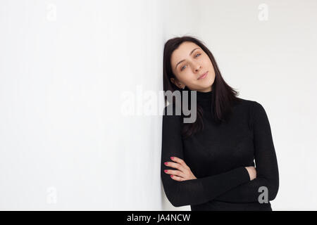 A girl in black clothes is standing by the white wall. She looks confidently forward, arms crossed by her chest Stock Photo