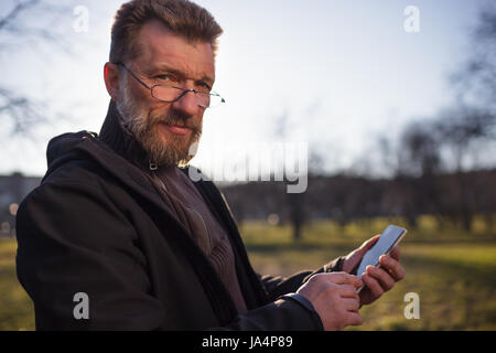Portrait of mature man with beard in park. He hold phone in hands Stock Photo