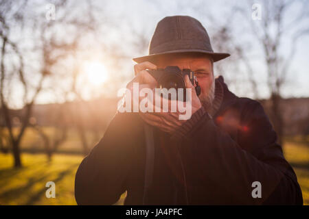 A mature man in a hat takes pictures on a mirrorless camera in the street. Hobby of photography. New trends in photographic technique. Stock Photo