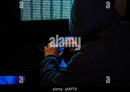 The man in the hood checks the malicious code on the keyboard. Cybercrime through the Internet. Stock Photo