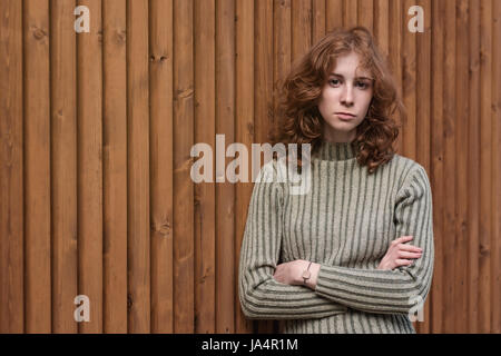 A beautiful red-haired girl in a green sweater is standing by the wooden wall. She crosses her arms and looks severely at the camera. Stock Photo