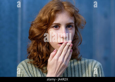 A red-haired, curly-haired girl in a green pullover frightened her hand over her mouth. She is surprised and shocked by something. Stock Photo