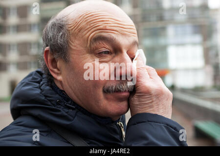 An elderly man with a mustache and bald head wipes tears in his eyes from the wind Stock Photo