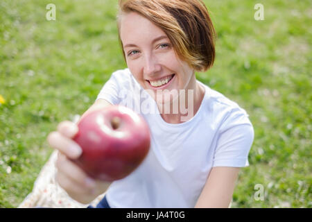Very beautiful caucasian model eating red apple in the Park. Outdoors portrait of pretty young girl sitting on green grass Stock Photo