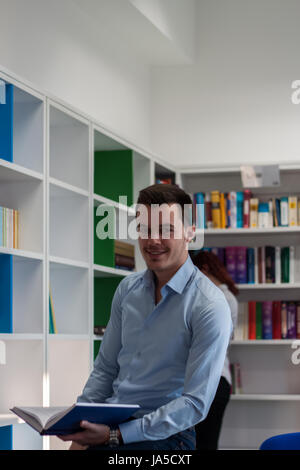 Handsome brunette student guy sitting on the desk and looking far away while having a book in his hands in the library. Bookshelf behind him Stock Photo