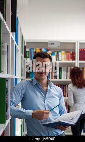 Handsome brunette student guy sitting on the desk and looking far away while having a book in his hands in the library. Bookshelf behind him Stock Photo
