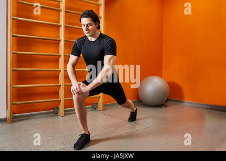 Strong young sportsman in the black sportwear stretching legs before gym workout. Stock Photo