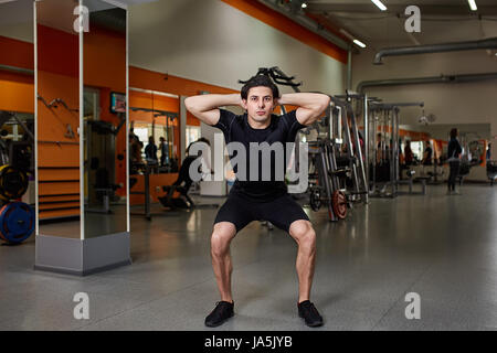 Portrait of young sportsman in the black sportwear while making squat in gym. Stock Photo