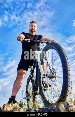 Bottom view of young cyclist standing with bike on rocks against blue sky. Stock Photo