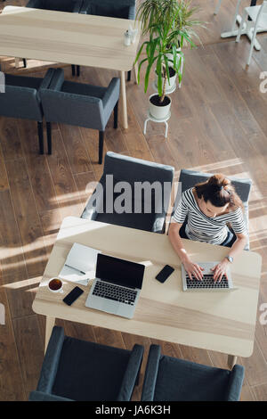 Overhead view or top view of a beautiful woman sitting at a table and working on her laptop. The sun casts graphics shadows on the wood floor. Stock Photo