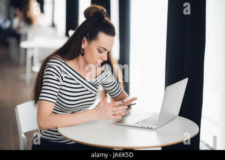 Young woman is sitting in a cafe and working during lunch time because of the deadline on her phone and laptop. Stock Photo
