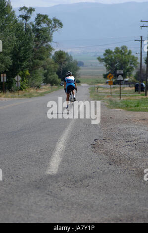 Woman on a road bike rides by herself on a long, straight road in rural Utah. Girl on a bicycle riding on the street in Spring City, UT. Stock Photo