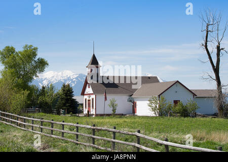 A country school house painted white with red trim, a flag and basketball hoop. A wooden fence is in foreground, rugged mountains in the background. Stock Photo