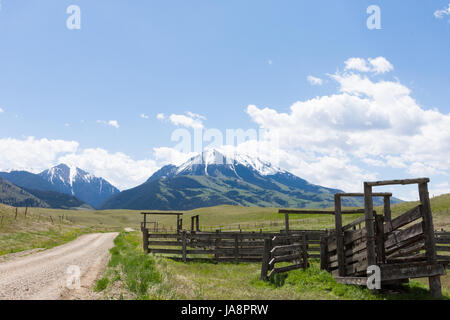 Close up of a old wooden corral in the foreground with a gravel road leading to rugged, snow capped mountains in the background. Stock Photo