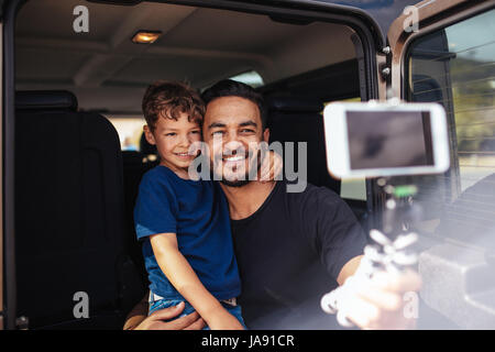 Handsome young dad and his cute little son sitting on back of the car making selfie using a smart phone. Father and son on road trip taking selfie. Stock Photo