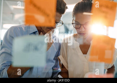 Two business professionals standing behind the glass wall with sticky notes and discussing. Colleagues brainstorming on new business ideas in office. Stock Photo
