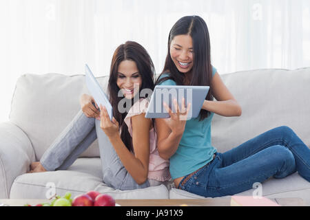 Friends sitting back to back showing each other their tablet pcs at home on the couch Stock Photo