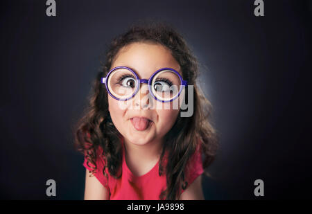 Caricature of a brunette curly six years girl with glasses sticking out tongue. Studio shot. Black background. Stock Photo
