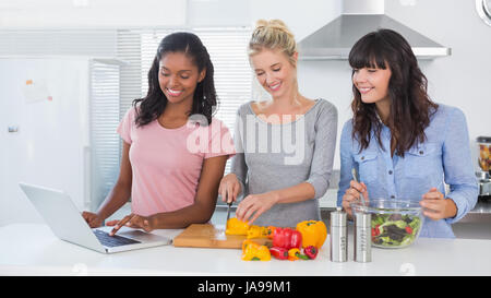 Happy friends making salad and using laptop for recipe at home in kitchen Stock Photo