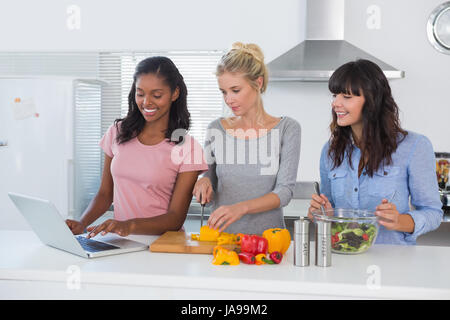 Smiling friends making salad and using laptop for recipe at home in kitchen Stock Photo