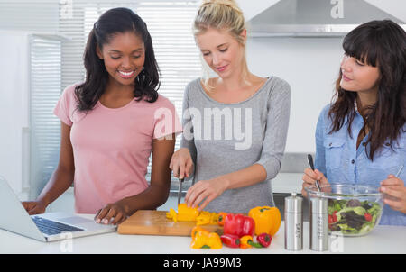 Cheerful friends making salad and using laptop for recipe at home in kitchen Stock Photo