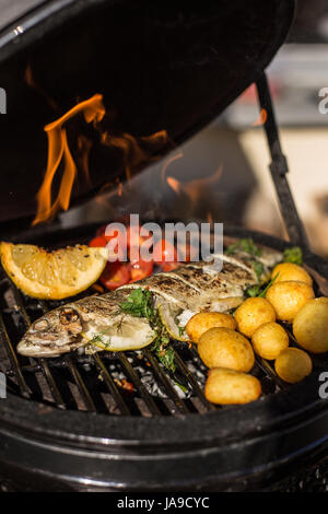 Delicious rainbow trout fish with tomatoes, potatoes and lemon cooking on hot flaming grill. Barbecue. Restaurant Stock Photo
